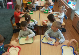 Dzieci siedzą przy stolikach rysują wzór palcem na tackach z kaszą manną.
